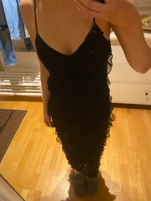 Svart långklänning med volanger - Säljer en svart långklänning med tunna axelband och v-ringning. Klänningen har volangdetaljer och en slit på ena benet