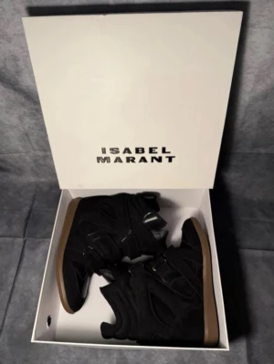  Isabel Marant svarta  - Säljer ett par svarta sneakers från Isabel Marant med högt skaft och platt brun sula. Skorna har en cool design med paneler i mocka och syntet, samt snörning och kardborreband för extra stil. Perfekta för dig som gillar statement-skor med edge.