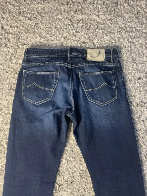 Jacob Cohën mörkblå jeans - Snygga mörkblå jeans från Jacob Cohën med kontrasterande vita sömmar och klassisk femficksdesign. Jeansen har en tydlig logotyp på bakfickan och patch i ljus färg vid linningen. Materialet är jeans och passformen är normal med rak siluett.