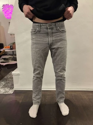 Grå jeans från Jack & Jones - Säljer ett par grå jeans från Jack & Jones med klassisk femficksdesign och ljus tvätt. Jeansen har rak passform och normal midja, tillverkade i bomullsmaterial. Snygga detaljer med märkets patch bak i midjan och diskreta sömmar. Modellen är 175 cm, vikt 68 kg