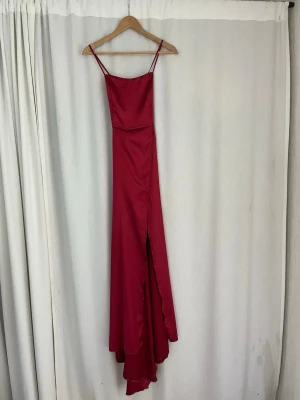 Vinröd långklänning med snörning - Säljer en vinröd långklänning i satin med smala axelband och snygg slits framtill. Använd en gång. Klänningen har öppen rygg med dekorativ snörning och en elegant, figurnära passform. Perfekt för bal eller student! Jag är 170 och har ca 5cm klackar, passade bra i längden. 