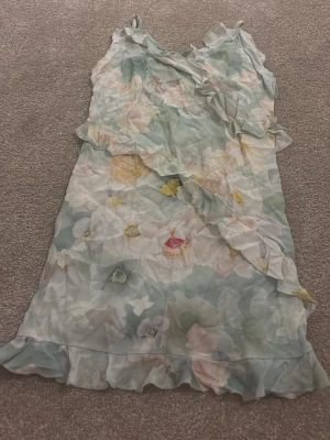 Blommig kortklänning med volang - Säljer en söt kortklänning från H&M i storlek XS. Klänningen har ett blommigt mönster i ljusa pastellfärger som blå, grön, gul och rosa. Den är ärmlös med tunna axelband och har volangdetaljer längs kanten och omlott framtill. Perfekt för sommarens alla tillfällen!