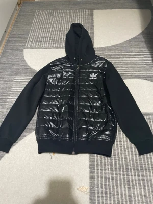 Svart pufferjacka från Adidas, strl L - Svart pufferjacka från Adidas med glansig quiltad framsida och mjuka ärmar. Jackan har huva med snörning, dragkedja och två fickor. Adidas-logga i vitt på bröstet. Perfekt för dig som gillar streetwear och vill ha en snygg jacka med coola detaljer. Det är storlek L men det motstår som storlek S. Den funkar som både jacka och kofta.