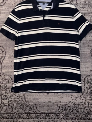 Tommy Hilfiger pikétröja - Säljer en klassisk pikétröja från Tommy Hilfiger i marinblått och vitt med horisontella ränder. Tröjan har korta ärmar, krage med ljusblå insida och liten logga på bröstet. Perfekt för en chill och stilren look.