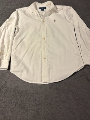 Vit långärmad skjorta från Ralph Lauren - Vit skjorta från Ralph Lauren med klassisk krage och broderad logga på bröstet. Skjortan har långa ärmar, knappar framtill och är gjord i mjuk bomull. Perfekt för dig som gillar stilrena och tidlösa plagg med snygga detaljer. Storlek 14 år i barn. Inga defekter