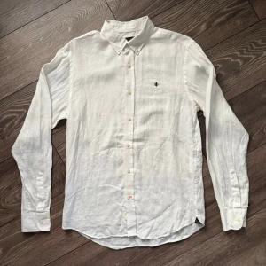 Vit linneskjorta från Morris - Säljer en stilren vit skjorta från Morris i ren linne. Skjortan har klassisk krage, knappar hela vägen och ett broderat emblem på bröstet. Långärmad och perfekt för varmare dagar. Passar dig som gillar enkel och fräsch stil. Lite missfärgad i armhålorna, därav priset