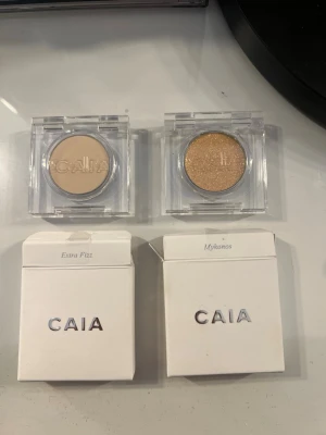 CAIA ögonskuggor Extra Fizz & Mykonos - Två singelögonskuggor från CAIA i transparenta fyrkantiga förpackningar. Extra Fizz är en matt beige nyans och Mykonos är en skimrande guldig ton. Perfekt för att skapa både naturliga och glammiga looks. Kommer med originalkartong. Helt orörda!