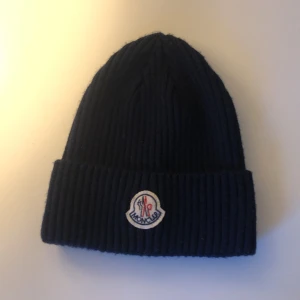 Mörkblå stickad Moncler mössa - Säljer en mörkblå ribbstickad mössa från Moncler med klassisk logotyp framtill. Mössan har bred uppvikt kant och är tillverkad i mjukt stickat material som håller dig varm och snygg under kyliga dagar.