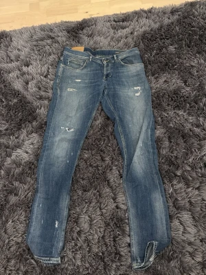 Blåa dondup jeans med slitningar - Säljer ett par blåa dondup  jeans med snygga slitningar och tvättade detaljer. Modellen har klassisk femficksdesign, gul kontrastsöm och logga på bakfickan. Jeansen är tillverkade i jeansmaterial och har en slim passform som sitter schysst på benen. Jeansen har blivit lagade i skrevet vilket visas på bild 4, vilket inte syns vid användning!