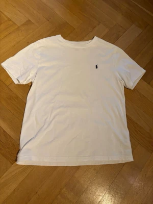Vit t-shirt från Polo Ralph Lauren - Vit t-shirt från Polo Ralph Lauren med klassisk rund hals och korta ärmar. Diskret Polo-logga broderad på bröstet. Tillverkad i mjuk bomull som känns skön mot huden. Perfekt för en clean och enkel stil.