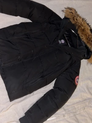 Svart Canada Goose dunjacka med huva - Dragkedjan har lossnat, men man kan fixa det. Storlek S, passar även M, päls ingår. Svart dunjacka från Canada Goose med huva och klassisk logopatch på ärmen. Jackan har långa ärmar, stora fickor och knappar framtill. Materialet är slitstark polyester och dunfyllning, perfekt för kalla dagar. Snygg, stilren och riktigt varm.
