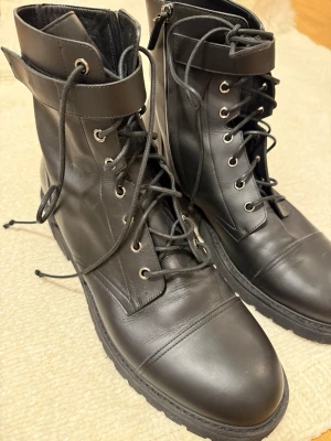 Svarta boots i skinn med snörning - Säljer ett par svarta boots i skinn med grov sula, snörning och dragkedja på sidan. De har rund tå och en bred rem upptill med metallhål. Perfekta för dig som gillar en tuff och stilren look.
