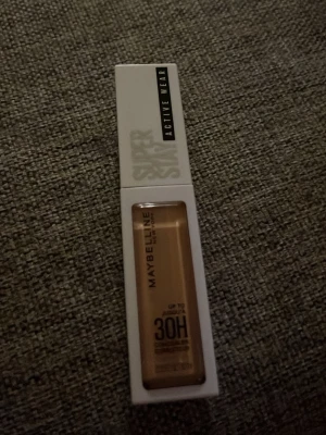 Maybelline Super Stay Concealer 25 - Maybelline Super Stay Active Wear Concealer i nyansen 25. Kommer i en smidig rektangulär tub med vit kork och transparent fönster som visar den beige färgen. Håller upp till 30 timmar och är perfekt för att täcka mörka ringar och ojämnheter. Helt ny 