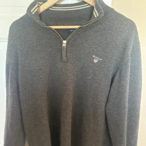 Grå half zip tröja från GANT - Säljer en grå långärmad tröja från GANT i superfin lammull. Tröjan har en half zip-dragkedja och ribbad krage med vita ränder. Klassisk GANT-logga broderad på bröstet. 