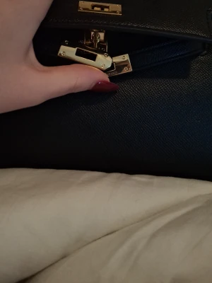 Svart handväska med gulddetaljer - Säljer en stilren svart handväska. exklusiva guldfärgade metalldetaljer och klassiskt lås framtill. Väskan är tillverkad i strukturerat skinn och har en elegant, boxig form med handtag upptill. Perfekt för dig som gillar lyxiga accessoarer och tidlös design.