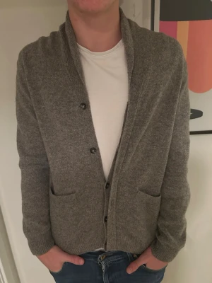 Grå kofta med knappar och fickor - Snygg grå kofta med ribbad krage och muddar, knäppning framtill med svarta knappar och två praktiska fickor. Perfekt att slänga på över en t-shirt för en chill och stilren look. Mjuk och skön stickad känsla.