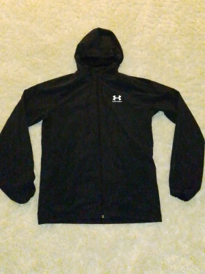 Svart vindjacka från Under Armour - Säljer en svart vindjacka från Under Armour med huva och dragkedja framtill. Jackan har lång ärm och en enkel, sportig design med logga på bröstet. Perfekt för utomhusaktiviteter och träning.