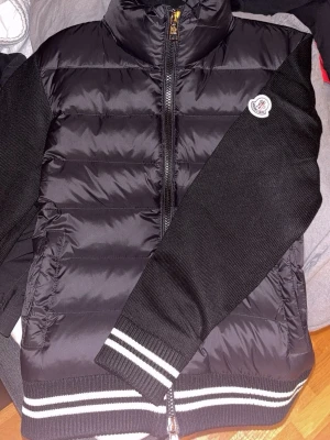Svart pufferjacka från Moncler - Svart pufferjacka från Moncler med stickade svarta ärmar och vita ränder på muddar och nederkant. Jackan kommer sitta riktigt fint på dig som har storlek S i jackor. Dragkedja framtill och Moncler-logga på ärmen. Materialet är en mix av polyester och stickad textil, perfekt för kyliga dagar. Hör av er för mer info om jackan. 