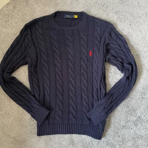 Mörkblå stickad tröja Polo Ralph Lauren - Kabelstickad mörkblå tröja från Polo Ralph Lauren med klassisk röd logga broderad på bröstet. Tröjan har rund halsringning, ribbade muddar och är långärmad. Storleken är S men upplevs som lite rymligare i passformen – perfekt om du gillar en mer relaxed/oversized look. Perfekt för dig som gillar preppy och stilren look.