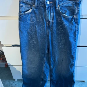 Blå jeans med mönster  - Snygga blå jeans från H&M Divided i storlek 34. Jeansen har ett unikt mönster framtill med text och snirkliga detaljer. Klassisk femficksmodell med raka ben och normal passform. Tillverkade i bomull för en skön känsla.