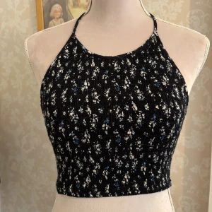 Blommig halterneck topp - Supersöt croppad halterneck topp från NLY Trend (köpt på nelly.com) i svart med små vita och blå blommor. Toppen har smockad struktur, knyt i nacken och baktill, och är ärmlös. Perfekt för varma sommardagar när du vill ha en chill och trendig look.