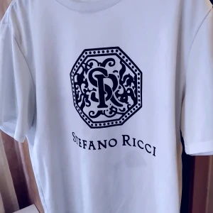 Vit t-shirt Stefano Ricci - Vit t-shirt från Stefano Ricci med stor svart logga och mönstrad emblem på bröstet. Klassisk rund hals och korta ärmar. T-shirten är gjord i mjuk bomull och har en clean, stilren vibe som passar perfekt till jeans eller shorts.