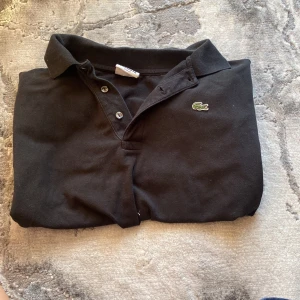 Svart Lacoste pikétröja i bomull - Svart pikétröja från Lacoste med klassisk krage och korta ärmar. Tröjan har tre knappar framtill och den ikoniska gröna krokodilloggan broderad på bröstet. Tillverkad i mjuk bomull som är skön mot huden. Perfekt för en chill och stilren look.