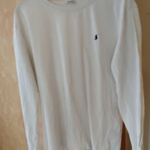 Vit långärmad tröja från Polo Ralph Lauren - Klassisk vit långärmad tröja från Polo Ralph Lauren med den ikoniska lilla broderade loggan på bröstet. Tröjan har rund halsringning och är gjord i mjuk bomull, perfekt för en clean och stilren look (Obs liten i storleken! Passar mer till M än L