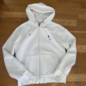 Vit hoodie från Polo Ralph Lauren - Snygg vit hoodie från Polo Ralph Lauren med dragkedja och klassisk broderad logga på bröstet. Tröjan har huva med snörning och stora fickor framtill. Perfekt för en clean och stilren look.