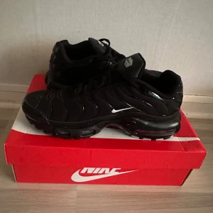 Nike Air Max Plus svarta sneakers - Nike Air Max Plus sneakers i helsvart med diskreta vita och färgglada detaljer på sidan. Skorna har en sportig vibe med synliga luftbubblor i sulan, mesh och syntetmaterial samt Tn-logga bak. Perfekta för dig som gillar streetstyle och vill sticka ut.