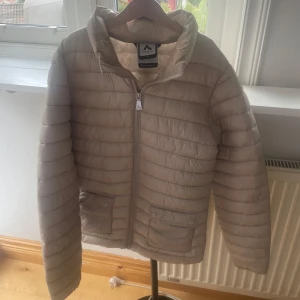 Beige pufferjacka från McKinley - Säljer en beige pufferjacka från McKinley med horisontella sömmar och två fickor framtill med knappdetaljer. Jackan har dragkedja hela vägen upp och en hög krage som skyddar mot vinden. Perfekt för kyliga dagar och enkel att matcha med allt.