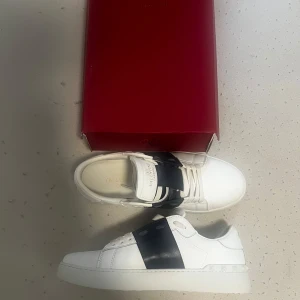Valentino Garavani Open sneakers vit/svart - Snygga Valentino Garavani Open sneakers i vitt skinn med bred svart läderdetalj över vristen. Klassisk rund tå, vita snören och ikoniska nitar på sulan. Perfekta för dig som vill ha en clean men lyxig look.