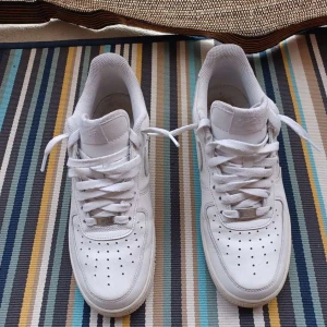 Vita Nike Air Force 1 sneakers - Klassiska vita Nike Air Force 1 sneakers med lågt skaft. Skorna har perforerad tå, rund tå och snörning. Tillverkade i skinn med vit sula och diskreta detaljer. Perfekta för en clean och stilren look.