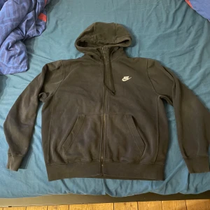 Svart Nike hoodie med dragkedja M - Svart hoodie från Nike i storlek M med vit logga på bröstet. Tröjan har huva med snörning, hel dragkedja och två stora fickor framtill. Perfekt för chill dagar eller när du vill ha en enkel och stilren look.