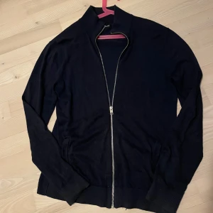 Svart kofta med dragkedja - En stilren mörkblå kofta från Jack & Jones med hel dragkedja framtill och ribbade muddar. Tröjan har lång ärm och två fickor på sidorna. Storlek S.  