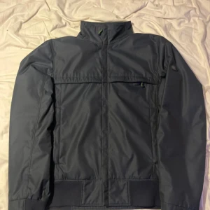 Svart vindjacka från Hugo Boss - Snygg svart vindjacka från Hugo Boss med diskret logga på ärmen och coola gröna detaljer. Jackan har hög krage, dragkedja och flera praktiska fickor. Insidan är fodrad med mönstrat tyg och jackan har en stilren, sportig look som passar perfekt till streetwear. I äffar kostar jackan 4990kr