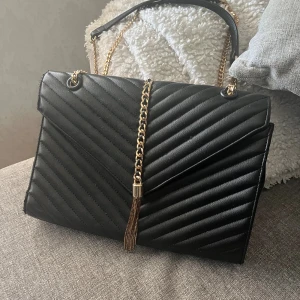 Svart axelväska  - Snygg svart axelväska med quiltat mönster och guldfärgad kedja som axelrem. Väskan har en dekorativ tofs framtill och ett praktiskt ytterfack på baksidan. Perfekt storlek för det viktigaste och ger en lyxig känsla till din outfit.