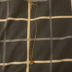 Guldigt halsband med rosettberlock - Snyggt halsband i guld med en unik berlock formad som en rosett. Kedjan är tunn och elegant, perfekt för att ge din outfit en extra touch. Rosetten har en cool 3D-effekt som verkligen sticker ut.