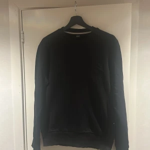 Svart sweatshirt från Hugo Boss - Svart sweatshirt från Hugo Boss med broderad logga framtill. Tröjan har rund halsringning, långa ärmar och ribbade muddar. Tillverkad i mjuk bomull för en skön och avslappnad stil.