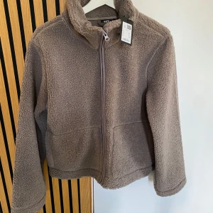 Brun  teddyjacka från 157, stl S - Mysig beige teddyjacka från 157 i storlek S. Jackan har hög krage, hel dragkedja framtill och är tillverkad i fluffigt teddy-material. Perfekt för kyliga dagar när du vill vara både varm och snygg. Enkel och stilren design som passar till det mesta. ALDRIG ANVÄND MED PRISLAPPEN KVAR 