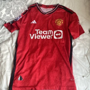 Manchester United hemmatröja Adidas - Manchester Uniteds officiella hemmatröja från Adidas med TeamViewer-tryck på bröstet och Premier League-märke på ärmen. Röd med svarta detaljer och subtilt grafiskt mönster. Klassisk rund hals och korta ärmar. Tillverkad i lätt och ventilerande funktionsmaterial.