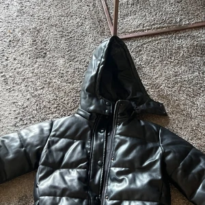 Svart pufferjacka i skinnimitation - Säljer en svart pufferjacka med huva och dragkedja. Jackan är quiltad och har en glansig yta i skinnimitation, perfekt för kalla dagar. Stängs med både dragkedja och tryckknappar framtill. Snygg och trendig modell som håller dig varm. Köptes för 1000- säljer för 600