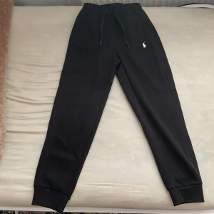 Svarta mjukisbyxor Polo Ralph Lauren S - Svarta mjukisbyxor från Polo Ralph Lauren med dragsko i midjan och broderad vit logga på vänster lår. Byxorna har muddar vid bensluten och är tillverkade i mjukt material för maximal komfort. Perfekta för chill dagar eller när du vill ha en avslappnad stil.