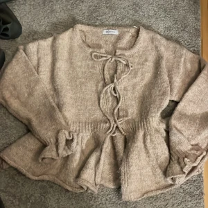 Beige stickad kofta med snörning - Superfin beige stickad kofta från JUSDEPOM med snörning framtill och volanger nedtill. Koftan har långa ärmar med volanger nedtill.