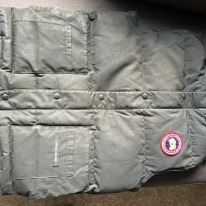Svart väst från Canada Goose - Snygg mörkgrå dunväst från Canada Goose med klassisk logopatch på bröstet. Västen har två stora fickor framtill, tryckknappar och quiltad design. Perfekt för kalla dagar när du vill ha en clean och stilren look. 
