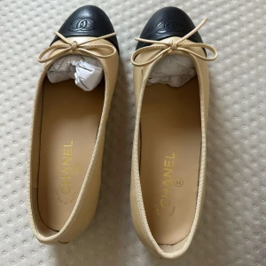 Chanel ballerinaskor beige & svart skinn - Klassiska ballerinaskor från Chanel i beige skinn med svart tå och ikonisk CC-logga. Söt rosett framtill och rund tå. Lyxig och tidlös design, perfekt för dig som gillar eleganta detaljer och premiumkänsla.
