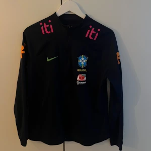 Nike Brasil fotbollströja marinblå - Svart Nike jacka med Brasil CBF-logga och sponsormärken i färg. Långärmad med dragkedja framtill och broderade detaljer. Perfekt för dig som älskar fotboll och vill ha en sportig look med officiell landslagsstil. (Använd av Pia Sundhage)