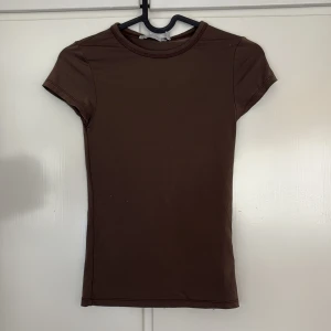 Brun kortärmad topp från 157 - En basic brun topp från 157 i storlek XS. Modellen är figurnära med rund halsringning och korta ärmar. Perfekt att styla med jeans eller kjol för en clean look. Tillverkad i mjukt material som känns skönt mot huden.