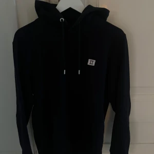 Mörkblå hoodie Les Deux - Mörkblå hoodie från Les Deux med huva och snörning, samt en liten broderad detalj med romerska siffran II på bröstet. Klassisk modell med långa ärmar och en avslappnad passform. Perfekt för dig som gillar stilrena och enkla plagg.