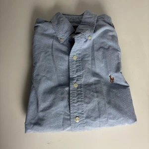 Blå skjorta från Ralph Lauren slim fit - Klassisk ljusblå skjorta från Ralph Lauren i slim fit-modell. Skjortan har långa ärmar, knappar framtill och broderad logga med polospelare på bröstet. Tillverkad i mjuk bomull och passar perfekt till en stilren look.
